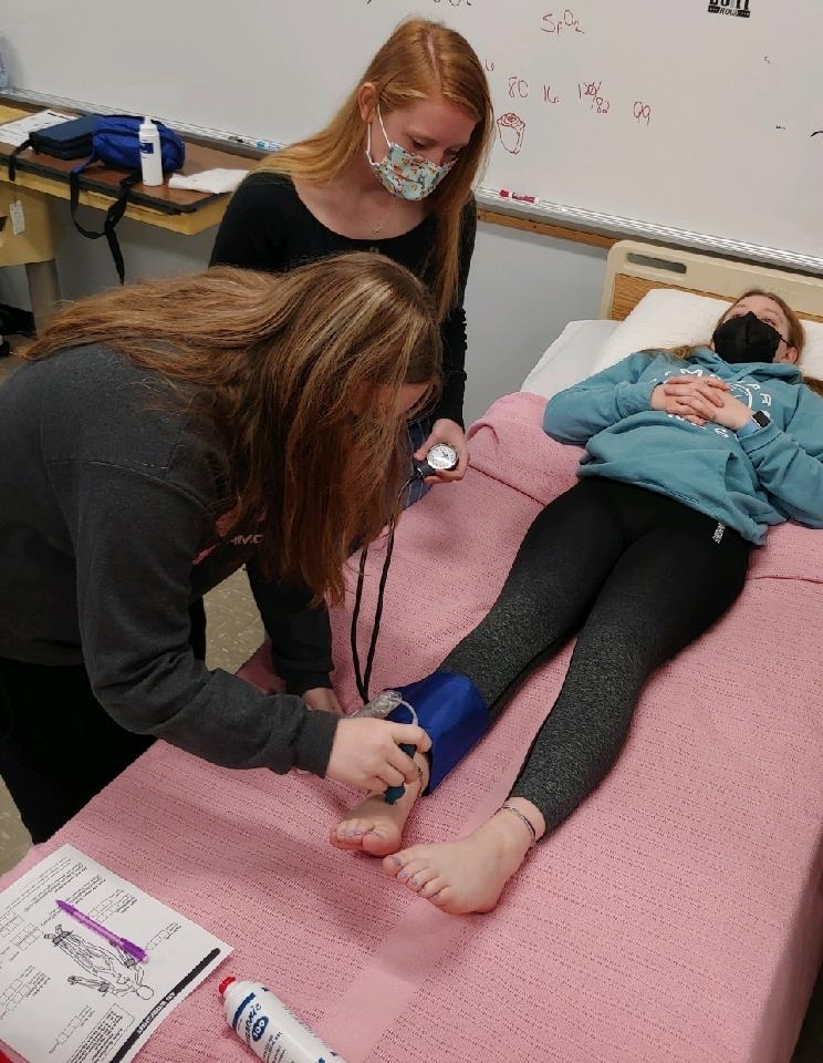 Two girls take a medical test on another girl lying in a hospital bed