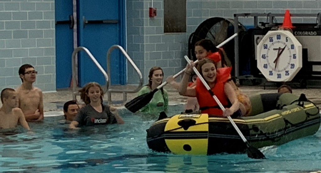 Students watch as 2 girls row a raft in the pool